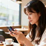 一位留着长发、约30岁的女性玩家头像，背景是明亮的咖啡厅，她拿着手机正在浏览游戏资讯。